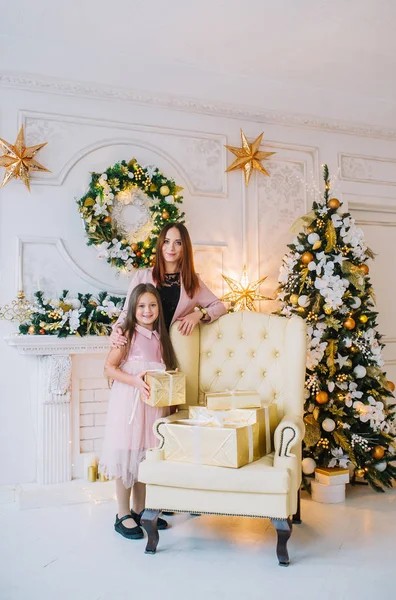 Niña en vestido rosa con su madre de pie cerca del sillón junto al árbol de Navidad en la habitación de Año Nuevo . — Foto de Stock