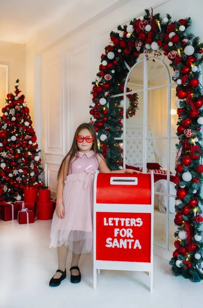 Engraçado menina em glases vermelhas fica perto de caixa de correio dentro de casa . — Fotografia de Stock