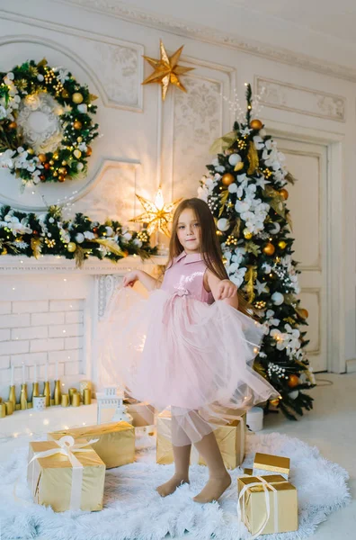 Una niña en un vestido rosa está bailando en la sala de estar cerca del árbol de Navidad con cajas de regalo — Foto de Stock