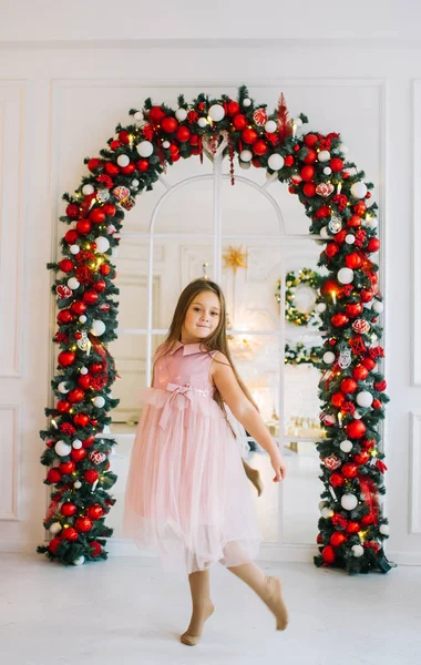 En liten flicka i en elegant rosa klänning dansar i ett rum mot bakgrund av en jul valv. — Stockfoto