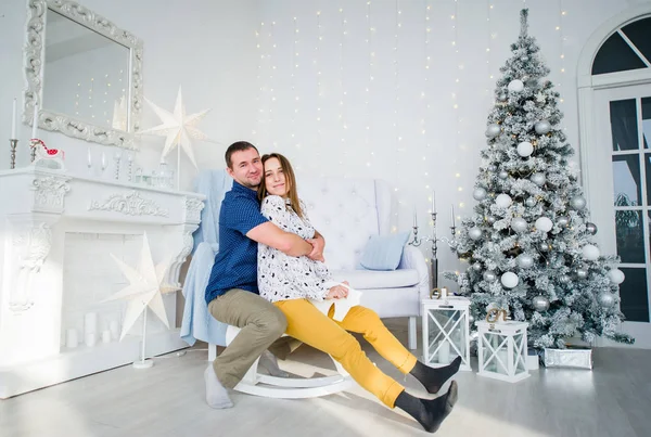 Guy en meisje in elkaars armen en zitten in de buurt van kerstboom in witte feestelijke kamer. Stockafbeelding