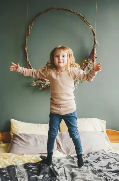 Little girl having fun and jumping on the bed in blue jeans and brown shirt on the background of a wall in decoration. — Stock Photo, Image