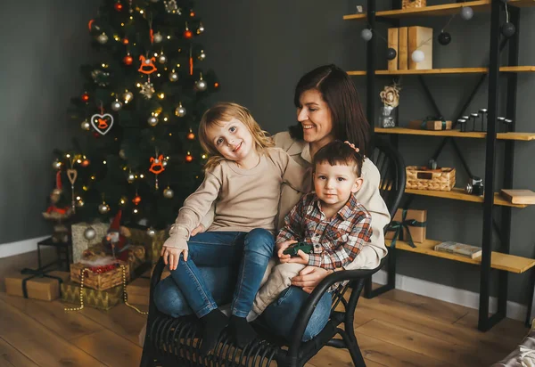 Genç beyaz bir anne ve çocukları süslü bir evin Noel ağacının yanında tahta bir sandalyede oturuyorlar.
