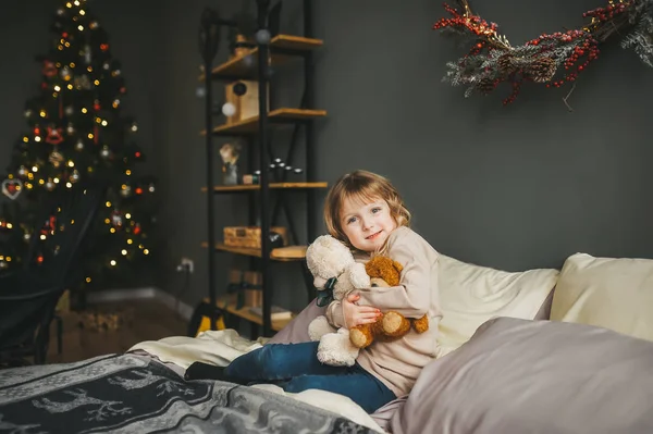 Glückliches kleines Mädchen mit Teddybär im Bett sitzend. Kind hat Spaß in der Weihnachtszeit. — Stockfoto