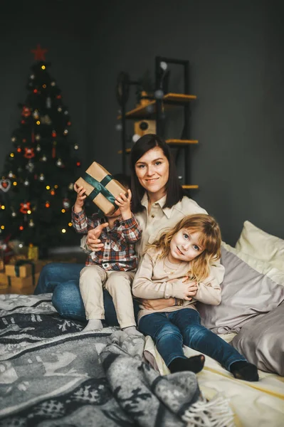 En glad ung mamma leker med sina söta små barn framför julgranen hemma. — Stockfoto