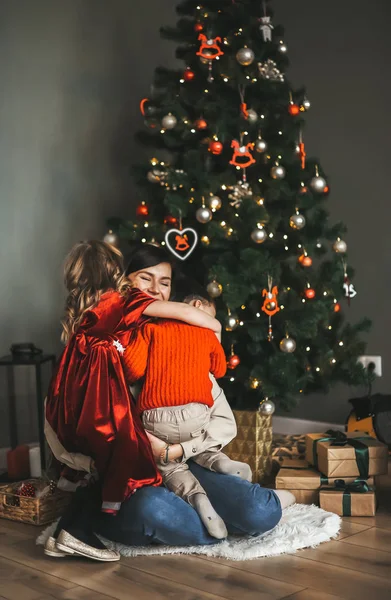 Una madre joven y feliz abrazándose con sus pequeños y lindos hijos frente al árbol de Navidad en su casa . — Foto de Stock