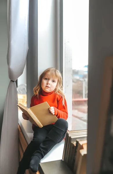 Feliz niña leyendo libro en casa mientras está sentado en el alféizar de la ventana — Foto de Stock