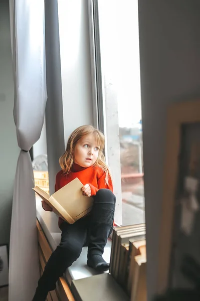 Niña con libro en las manos sentada en un alféizar de la ventana y mira a la calle — Foto de Stock