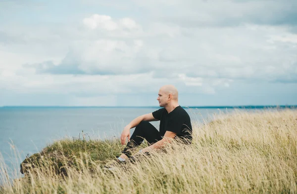 Ένας φαλακρός άνδρας με μαύρο πουκάμισο κάθεται στο γρασίδι στην άκρη της βραχώδους ακτής της Βαλτικής θάλασσας. — Φωτογραφία Αρχείου