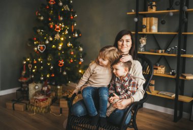 Anne çocuklarını kucaklıyor ve Noel ağacının yanındaki sandalyeye oturuyor.. 