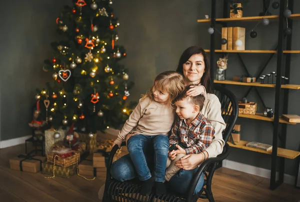 Modern omfamnar sina barn och sitter på stol nära julgranen. — Stockfoto