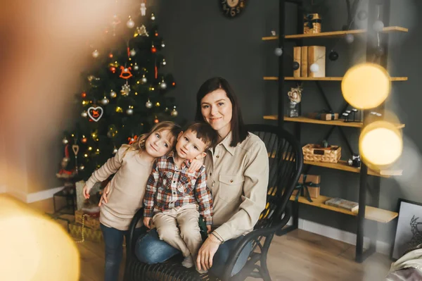 Mamma och hennes barn sitter på svart stol i festligt rum med julgran. — Stockfoto