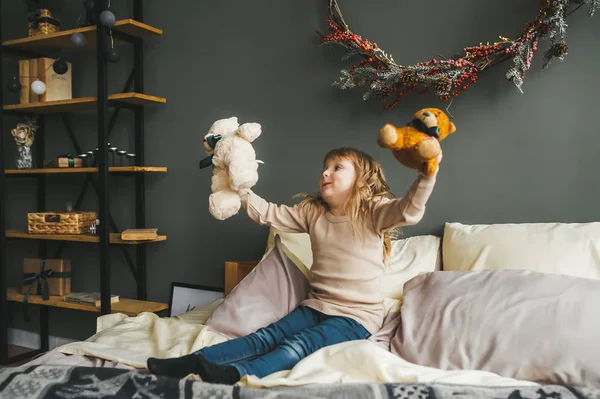 Hübsches kleines Mädchen, das mit einem Teddybärspielzeug spielt, das auf einem Bett neben einem Weihnachtsbaum sitzt. — Stockfoto