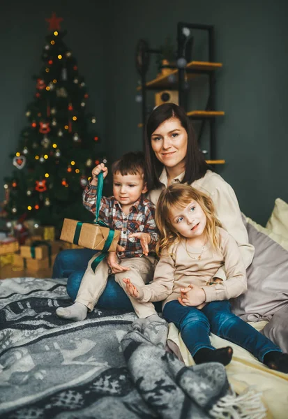 Lycklig familj med tre sittande på sängen i festligt rum med julgran tittar på kameran. — Stockfoto