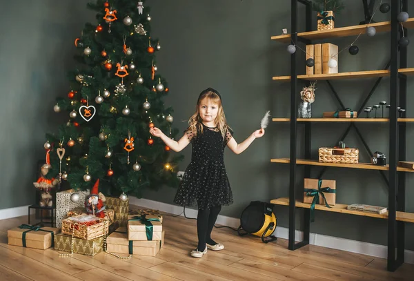En liten flicka med liten fjäder i händerna står i festligt hem rum nära julgran — Stockfoto