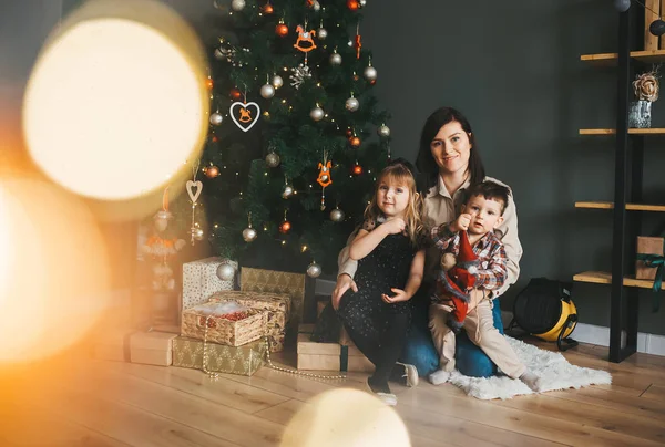 Familia sonriente con cajas de regalo sentadas en el suelo cerca del árbol de Navidad en el fondo de las luces — Foto de Stock