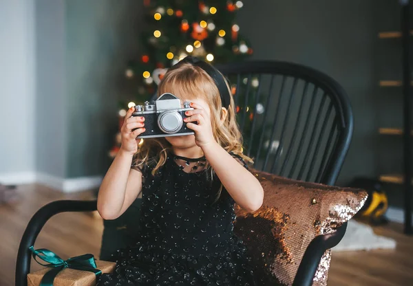 Un niño lindo te fotografía en una cámara retro sobre el fondo del árbol de Navidad con bolas . — Foto de Stock