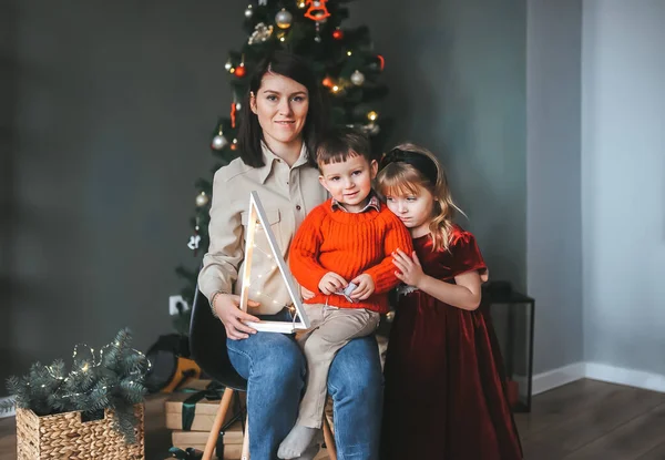 Retrato de la madre y dos niños sentados en una silla en casa cerca del árbol de Navidad . — Foto de Stock