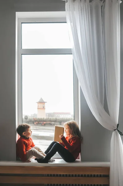 Niños leyendo libros sentados en el alféizar de la ventana en casa. Niño y niña leyendo a la luz del amanecer . — Foto de Stock