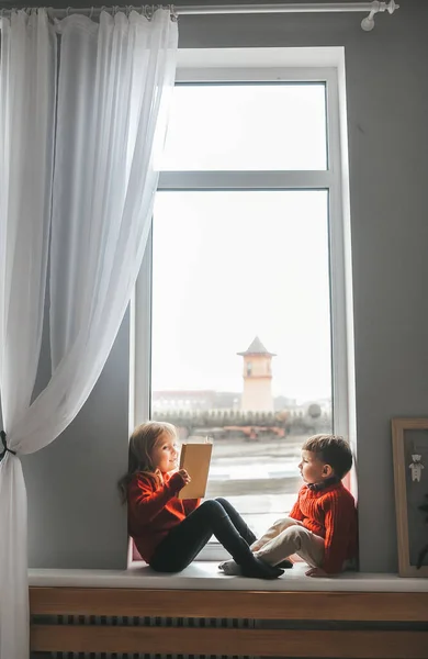 Niño y su hermana en casa jugando en el alféizar de la ventana y la lectura de libros . — Foto de Stock