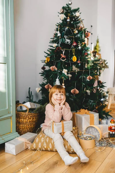 Leende liten flicka sitter under julgran med presenter och presenter i rummet. — Stockfoto