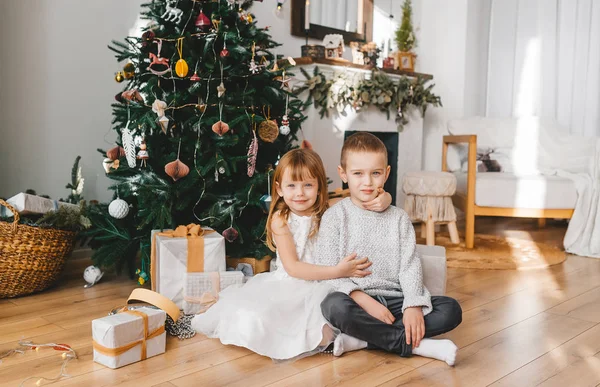 Retrato navideño de hermosos niños sonrientes sentados en el suelo con regalos bajo el árbol de Navidad . — Foto de Stock