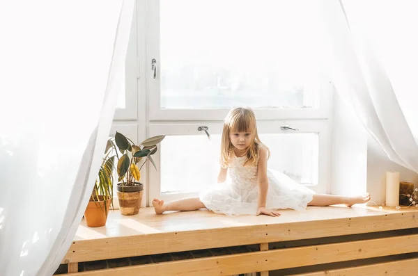 Cute little girl in beautiful dress is drop into the splits on a window sill. — Stock Photo, Image