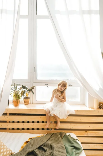 Linda niña en hermoso vestido está sentado en un alféizar de la ventana en la luz dormitorio soleado — Foto de Stock