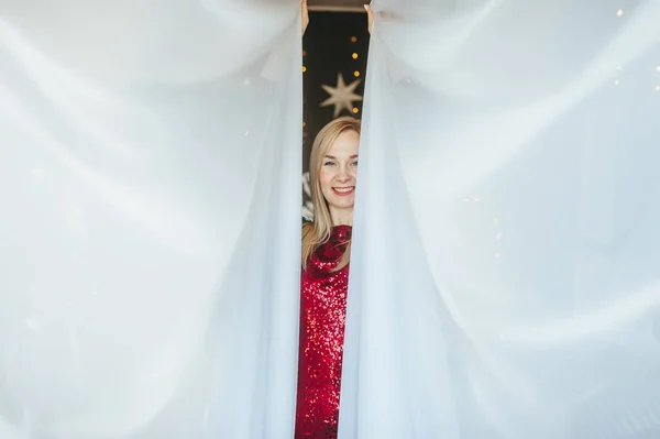 Блондинка в красном платье, стоящая в гостиной за шторами и улыбающаяся . — стоковое фото