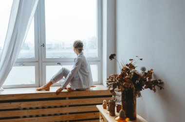 Resting and thinking woman in pajamas sitting on the window-sill at home.
