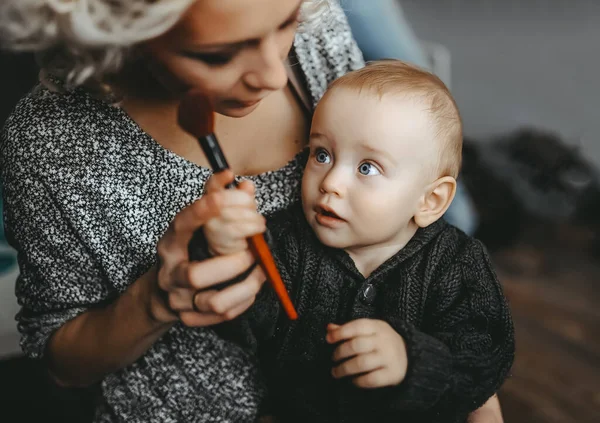 Glücklich lächelnder kleiner Junge blickt auf ihre schöne Mutter. Mutter schminkt sich mit Kosmetikpinsel. — Stockfoto