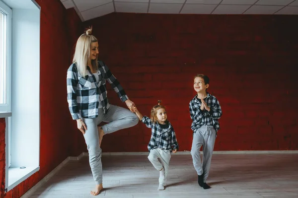 Madre joven haciendo ejercicios de yoga con sus dos hijos cerca de la ventana en casa . — Foto de Stock