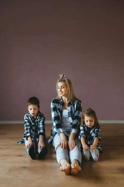 Two cute kids and her beautiful mother in identical clothes sitting on a wooden floor. — Stock Photo, Image