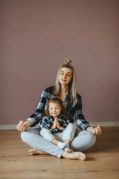 Young woman sitting on the floor in a living room with her little daughter in a lotus pose with closed eyes. — Stock Photo, Image