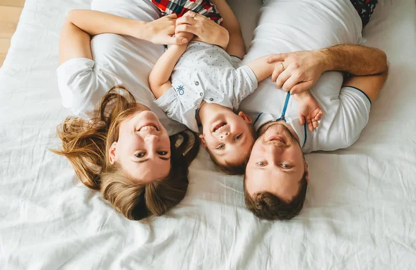 Ouders met zoon liggen samen op bed, wit beddengoed glimlachend kijkend naar de camera voelt zich gelukkig. Bovenaanzicht. Stockfoto