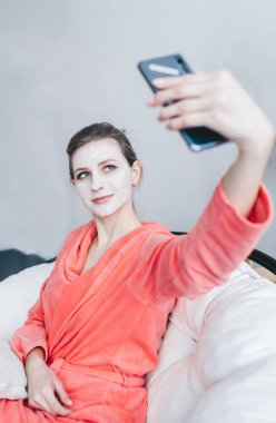 Kırmızı bornozlu güzel bir kadın, kanepede oturuyor, selfie çekiyor, evde güzellik prosedürleri sırasında dinleniyor..