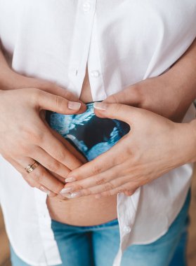 Karı ve kocanın ellerinin hamile karnına kalp şekli vermesi. Yüzler yok. Dikey görünüm