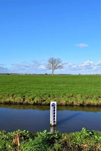 Rural scenery with water level meter in ditch and tree in meadow and blue sky with clouds