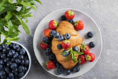 Fransız kahvaltısı, taze kruvasan, çilek ve yabanmersinli. Gri arkaplan, yeşil nane yaprakları. Fırın ürünleri