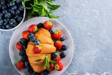Fransız kahvaltısı, taze kruvasan, çilek ve yabanmersinli. Gri arkaplan, yeşil nane yaprakları. Fırın ürünleri