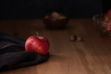 Koyu kahverengi kumaş üzerinde parlak kırmızı bir elma. Arka planda fındık ve karamsar bir ışıklandırma.