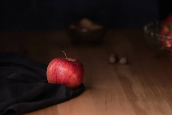 Koyu kahverengi kumaş üzerinde parlak kırmızı bir elma. Arka planda fındık ve karamsar bir ışıklandırma.