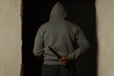 A man in a hood on his head holds a knife in his hand behind his back.