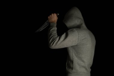 A man in a hood on a black background, brandishing with a knife in his hand. A criminal with a knife attacks from the dark.