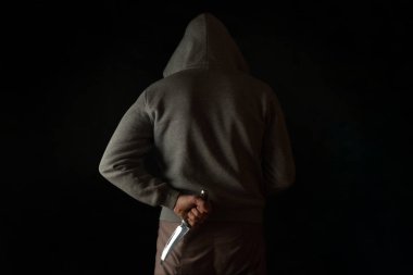 A man holds a knife in his hand behind his back, in a hood on a black background. Criminal with a knife hiding in the dark.