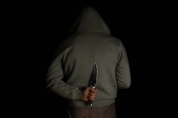 A man holds a knife in his hand behind his back, in a hood on a black background. Criminal with a knife hiding in the dark.