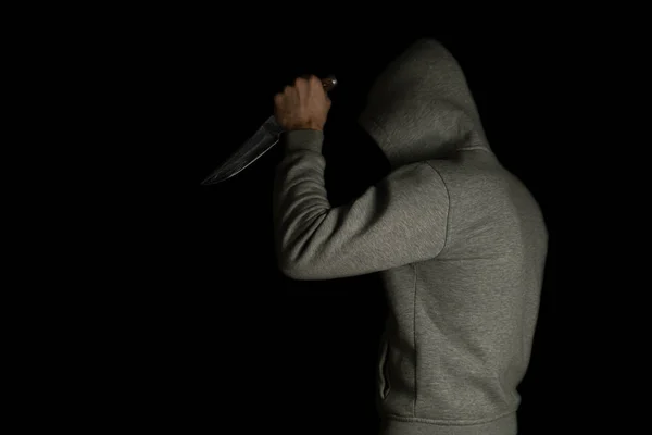 A man in a hood on a black background, brandishing with a knife in his hand. A criminal with a knife attacks from the dark.