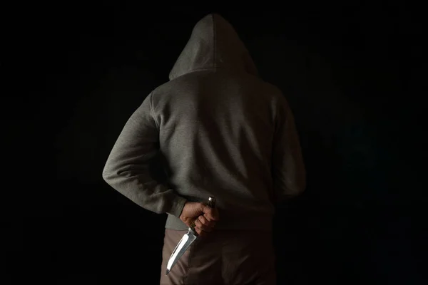 A man holds a knife in his hand behind his back, in a hood on a black background. Criminal with a knife hiding in the dark.