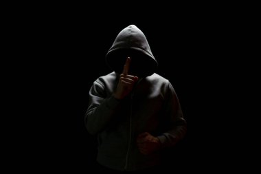 Kapüşonlu, siyah yüzlü bir adam karanlıktan çıkar ve işaret parmağını kaldırarak susmasını ister.