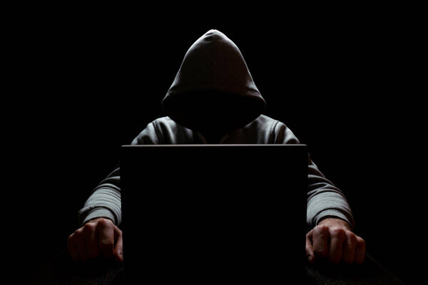 злобный компьютерный хакер в капюшоне сидит в темноте, руки протянуты, по бокам монитора ноутбука, избирательный фокус
